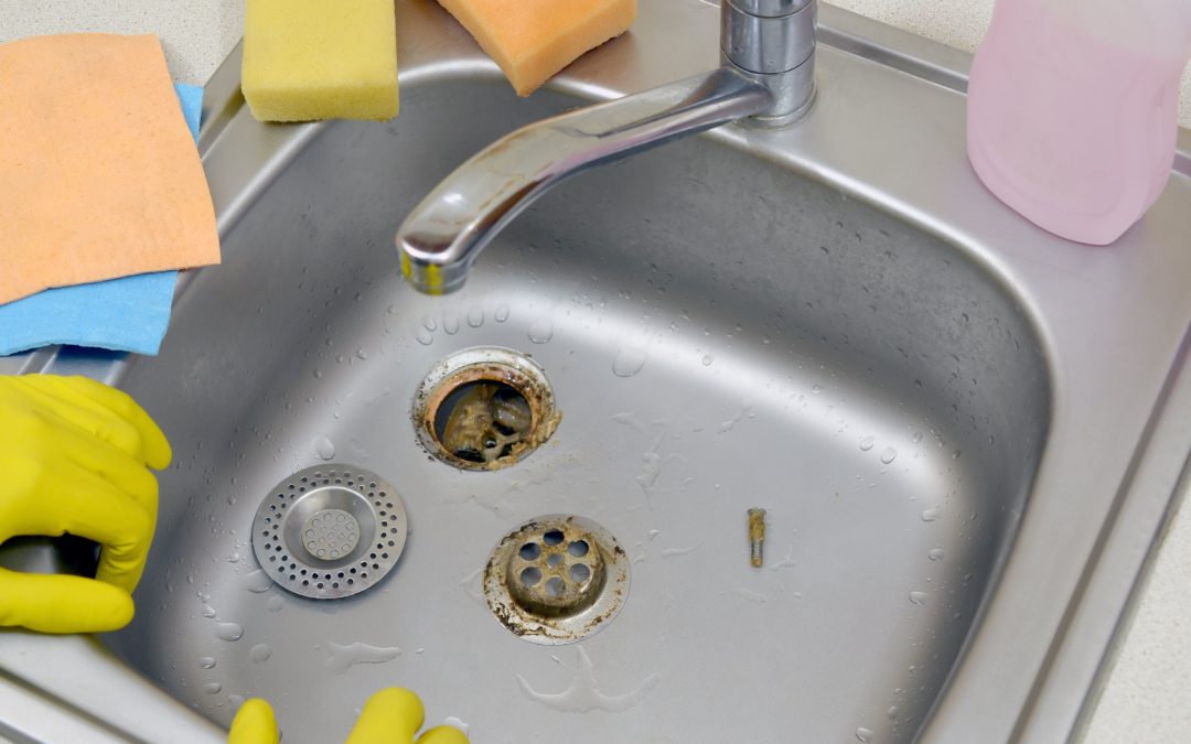 A clogged and broken kitchen sink drain. A plumber wearing yellow gloves is trying to fix it.